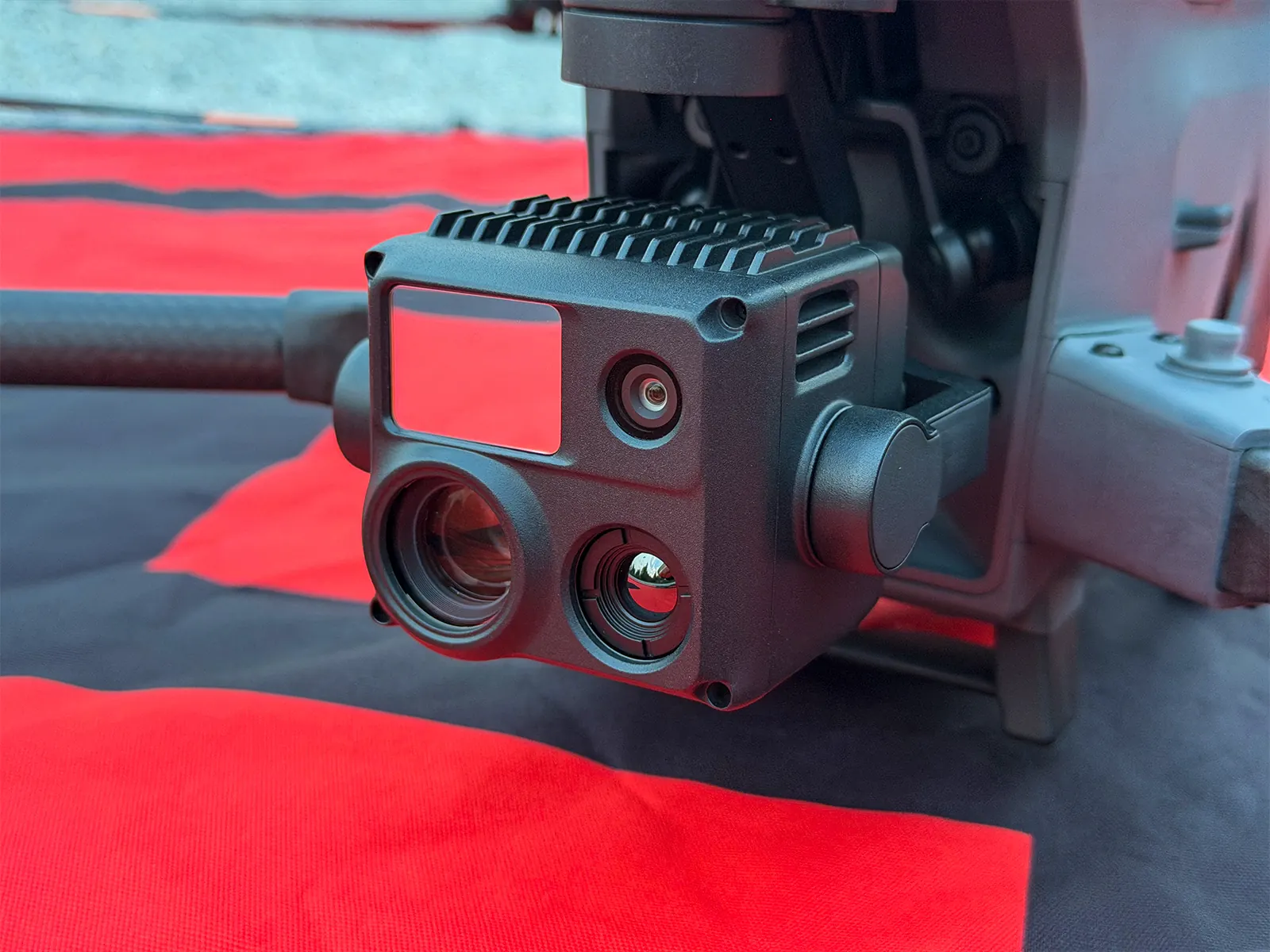 Close-up of the DJI Matrice 30T gimbal camera showing the thermal, zoom and wide lenses and laser rangefinder, on a red landing pad.
